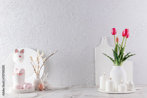 Fototapeta Naklejka Na Ścianę i Meble -  Kitchen background in light colors for the Easter holiday. Flowers and willows in vases, candles, Easter eggs and bunnies on a marble countertop.