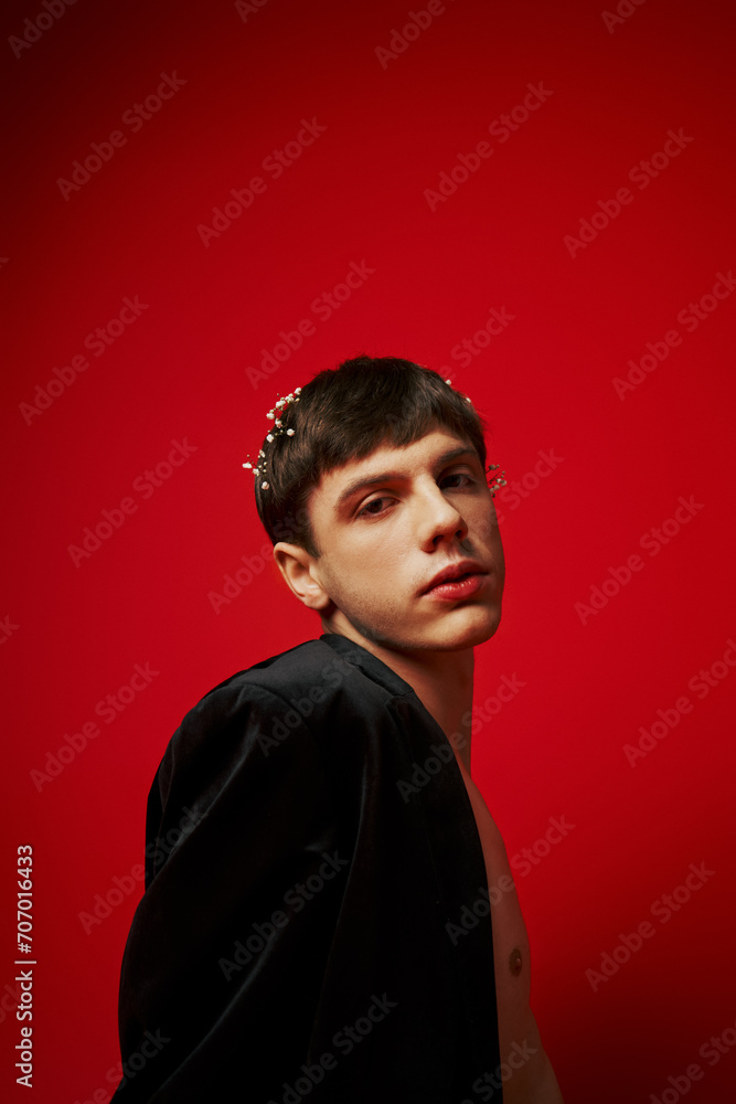 young fashionable and shirtless man in velvet blazer with flowers in hair posing on red background young fashionable and shirtless man in velvet blazer with flowers in hair posing on red background