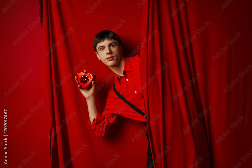 young man in red shirt with suspenders holding heart shaped gift box near curtain, 14 February