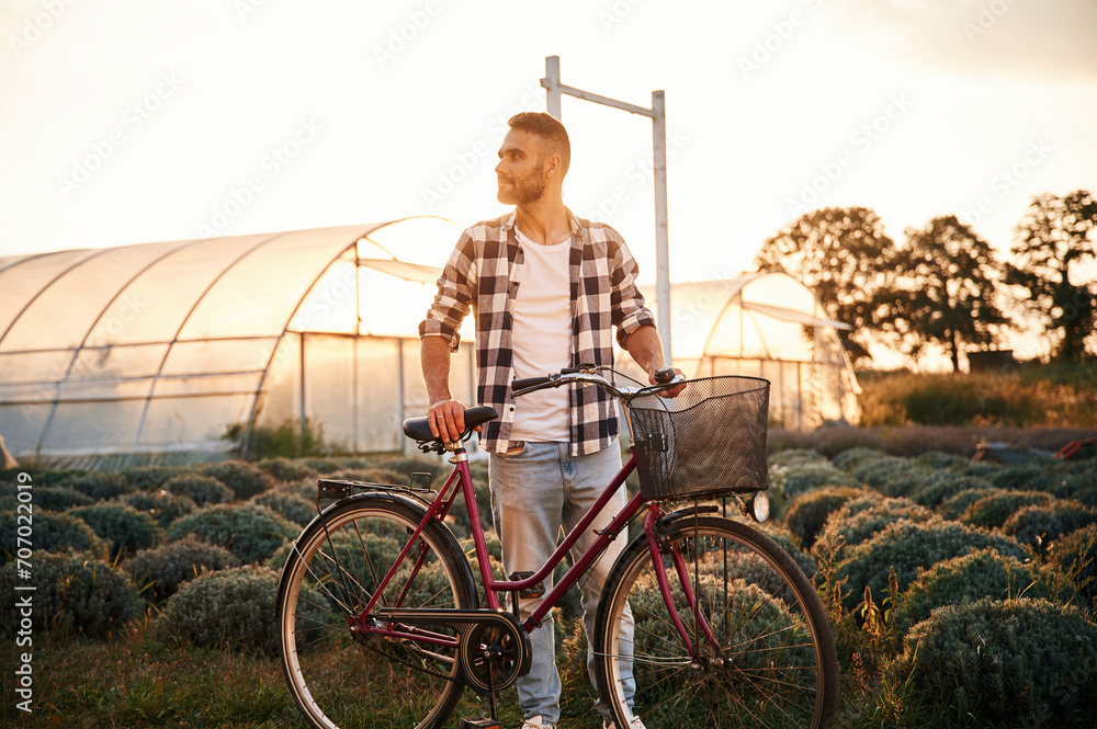 Obraz premium Standing, enjoying nature. Handsome man in casual clothes is with bicycle on the agricultural field near greenhouse