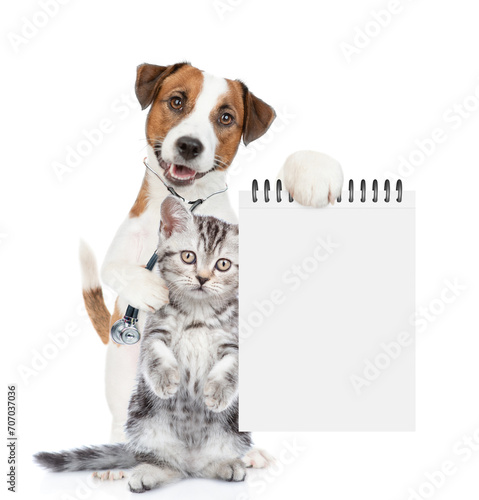 Smart jack russell terrier with stethoscope on his neck hugs tiny kitten and shows empty notebook. isolated on white background