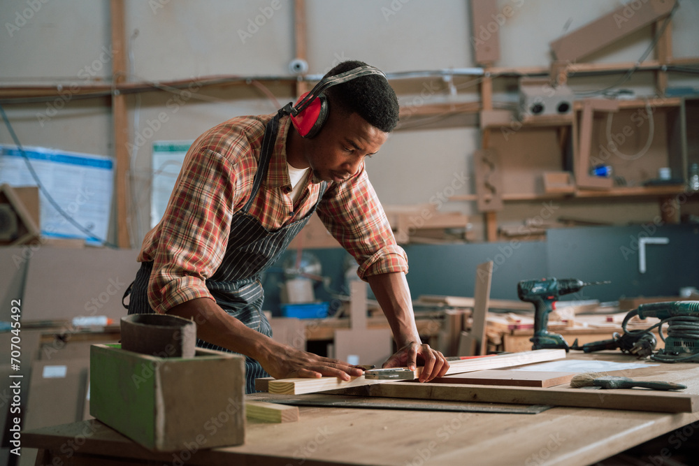 Concentrated African male wearing earmuffs measures piece of wood for carpentry project Concentrated African male wearing earmuffs measures piece of wood for carpentry project