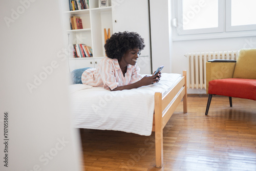 Happy young woman using smartphone while lying in bed at home