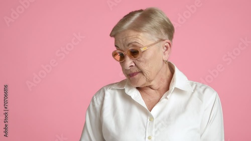 Medium shot of confused elegant wrinkled nice-looking lady of 60-70 years in white shirt turning glance away from camera, standing over pink background. High quality 4k footage