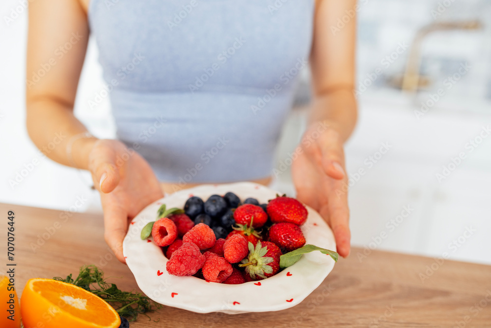 Close-up of female hands holding a ceramic bowl with healthy berries in the kitchen. Blueberries, raspberries and strawberries on a white plate. Close-up of female hands holding a ceramic bowl with healthy berries in the kitchen. Blueberries, raspberries and strawberries on a white plate.