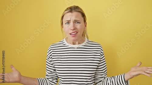 Bewildered blonde beauty in a striped tee, expressing 'don't know' gesture isolated on a sunny yellow backdrop – a case of adorable cluelessness or sheer indifference? photo