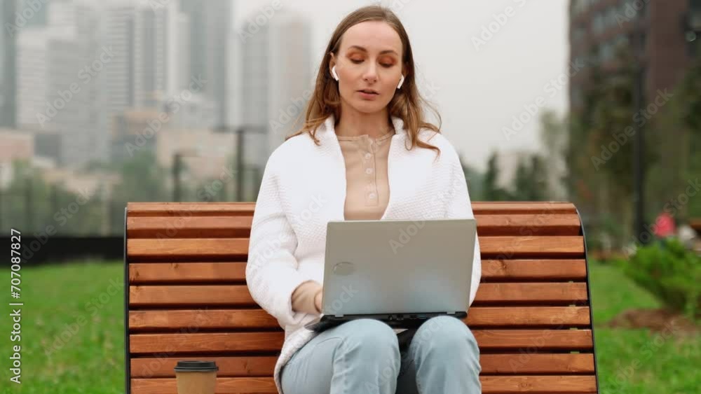 Happy woman wearing wireless headphones is talking online via video conference with a laptop while sitting on a bench outside. Happy woman wearing wireless headphones is talking online via video conference with a laptop while sitting on a bench outside.