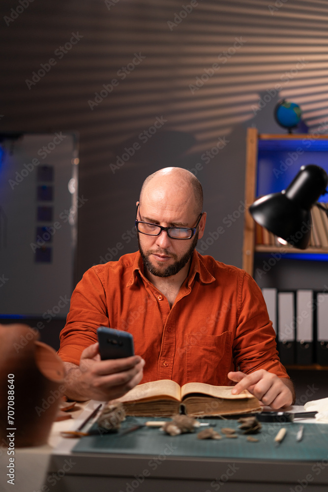Archeologist reading book and using mobile phone working late night in office or in research lab. Archaeology, paleontology and science concept. Archeologist reading book and using mobile phone working late night in office or in research lab. Archaeology, paleontology and science concept.