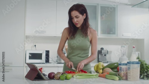 Medium shot of young Caucasian woman in green homewear making vegetable salad looking at recipe on digital tablet photo