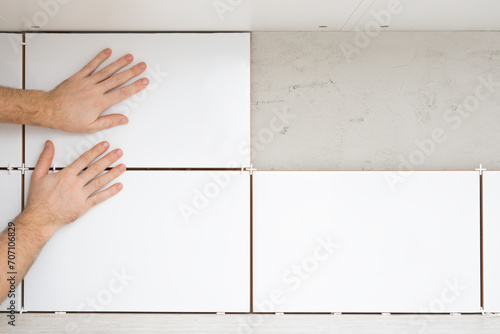 Young adult man hands gluing white ceramic tile on concrete wall between kitchen cabinets and table top. Closeup. Front view. Renovation process. Repair work of home. photo