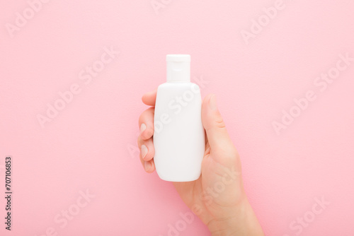 Young adult woman hand holding white plastic cream bottle on light pink table background. Pastel color. Care about clean and soft body skin. Daily female beauty product. Closeup. Top down view.
