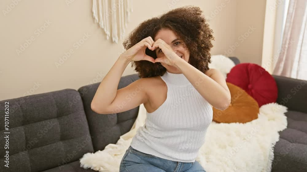 Cheerful hispanic woman with curly hair, sitting on living room sofa, showing a heart sign with hands. love-filled home filled with romance! Cheerful hispanic woman with curly hair, sitting on living room sofa, showing a heart sign with hands. love-filled home filled with romance!