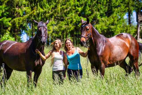 Two riders girls women next to each other in summer on a hay meadow in front of a fir forest.