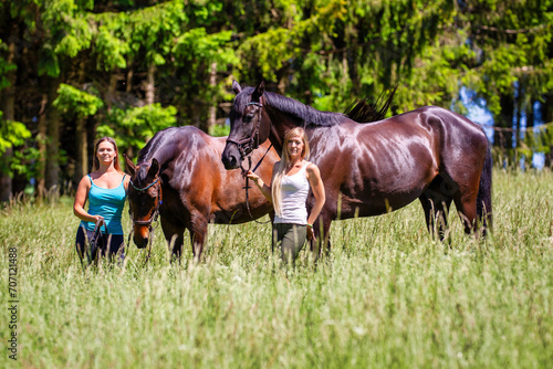 Two riders girls women next to each other in summer on a hay meadow in front of a fir forest.