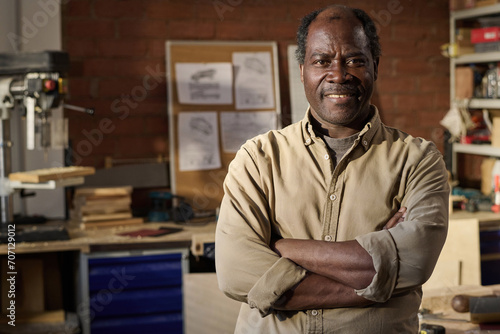 Waist up portrait of senior Black carpenter smiling at camera standing with arms crossed in workshop