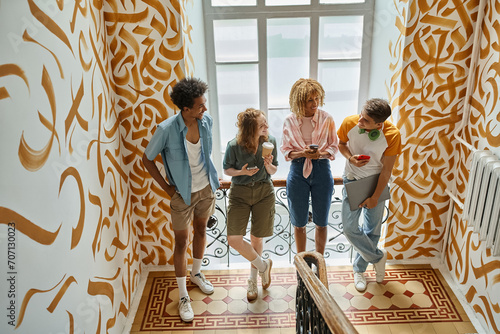 joyful multiethnic students with gadgets talking on staircase of modern youth hostel, travelers