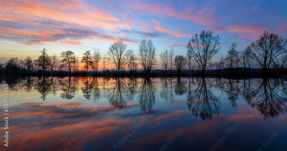 Colorful sunset panorama at Ruhr river near Iserlohn and Schwerte. Row of bare tree silhouettes with perfect reflection in calm water surface on a winters evening with pink clouds. Romantic atmosphere