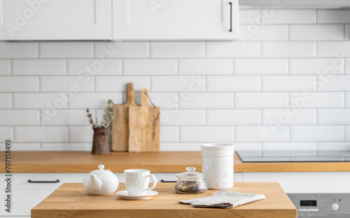 Wooden oak table with a cup of tea and a kettle in front of the kitchen with a white brick background.