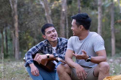Asian LGBTQ couple enjoying nature, camping with tents in the forest area by the river, playing guitar. © wichayada