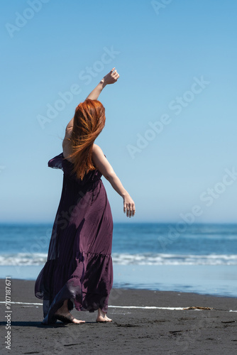 Redhead woman in dark puce long dress standing on sandy beach with hand raised, against backdrop of crystal blue sea on sunny summer day with clear sky. Woman is seen from rear in full length photo