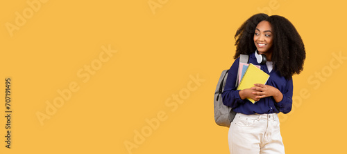 Smiling black student with notebooks looking to the side at free space on yellow background