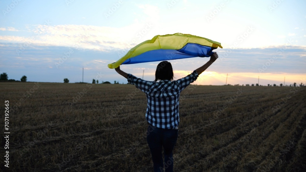 Ukrainian woman running with raised flag Ukraine above her head on wheat field at sunset. Lady jogging with national blue-yellow banner on barley meadow at sunrise. Victory against russian aggression.