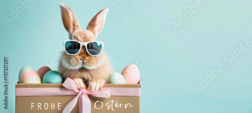 Frohe Ostern Konzept Feiertag Grußkarte mit deutschem Text - Cooler Osterhase, Kaninchen mit Sonnenbrille, sitzt in Geschenkbox mit Ostereiern, isoliert auf blauem Hintergrund photo