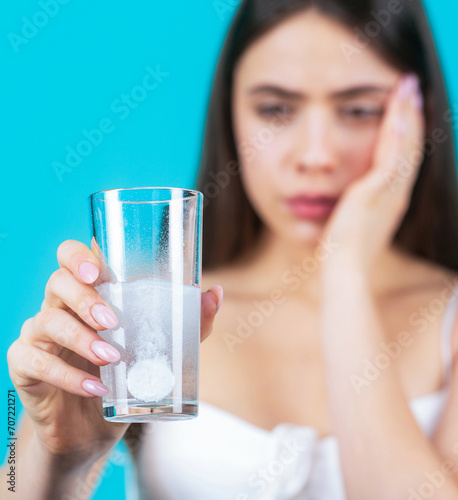 Woman taking drugs to releave headache. Brunette take some pills, holds glass of water, isolated on blue. Young woman taking pill against headache. Brunette taking a pill with a glass of water photo