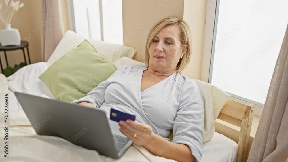Mature blonde woman shopping online with credit card in a cozy bedroom setting, expressing various emotions. Mature blonde woman shopping online with credit card in a cozy bedroom setting, expressing various emotions.