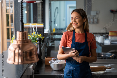 Successful woman baker wearing apron holding digital tablet pc looking through the window in bakery kitchen. Smiling blonde young woman with fintech device in pastry kitchen. photo