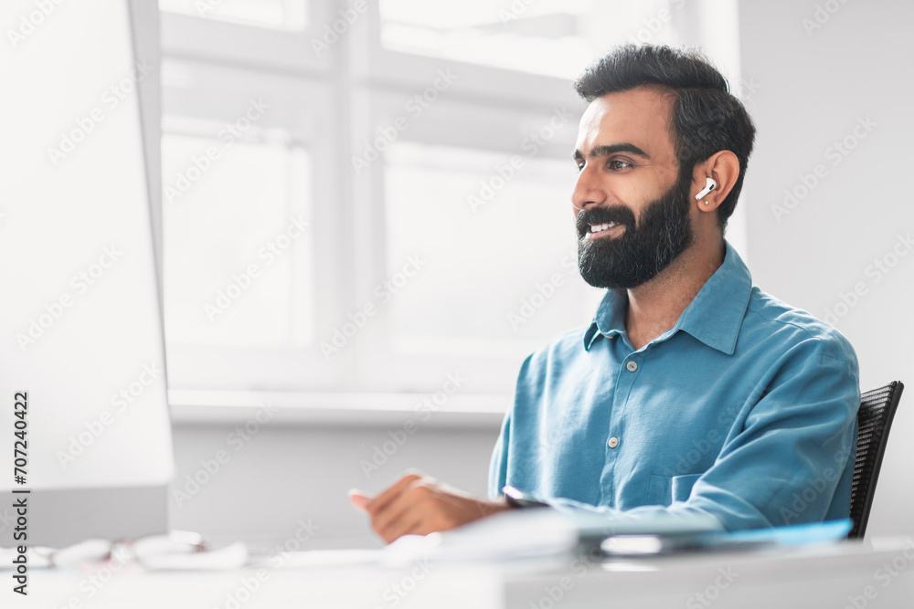 Obraz premium Smiling indian man with earbuds working at bright office desk