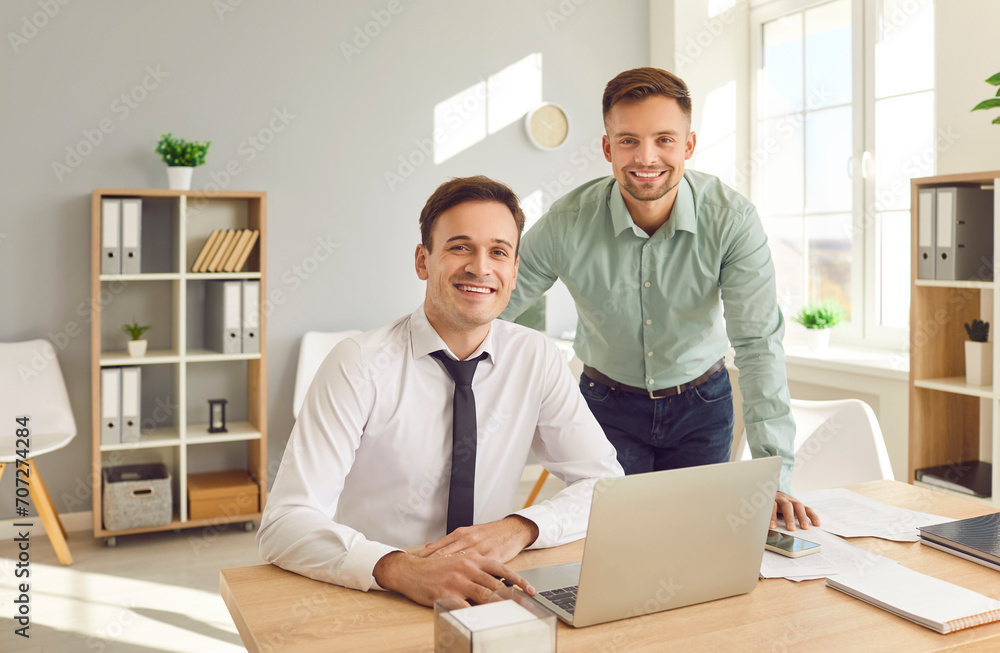 Portrait of a two happy smiling confident business people wearing shirts looking cheerful at camera at the workplace sitting at the desk. Company employees and male coworkers working in office. Portrait of a two happy smiling confident business people wearing shirts looking cheerful at camera at the workplace sitting at the desk. Company employees and male coworkers working in office.