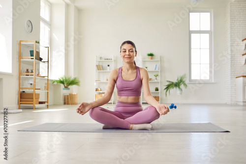 Woman doing yoga at home. Beautiful young girl in sportswear with closed eyes sitting on workout mat in her modern studio apartment, doing lotus pose, meditating, and relaxing