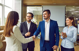 Group of business people in suits meeting in the office, making a deal and exchanging handshakes. Happy man and woman shake hands while colleagues applaud. Professional cooperation, teamwork concept