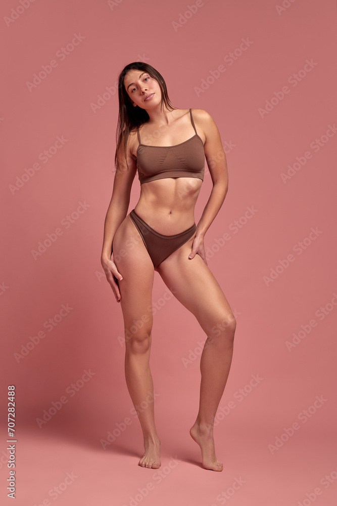 Full-length image of young beautiful girl with slim, healthy, fit body shape, posing in underwear against pink studio background. Concept of natural beauty, health and body care, wellness