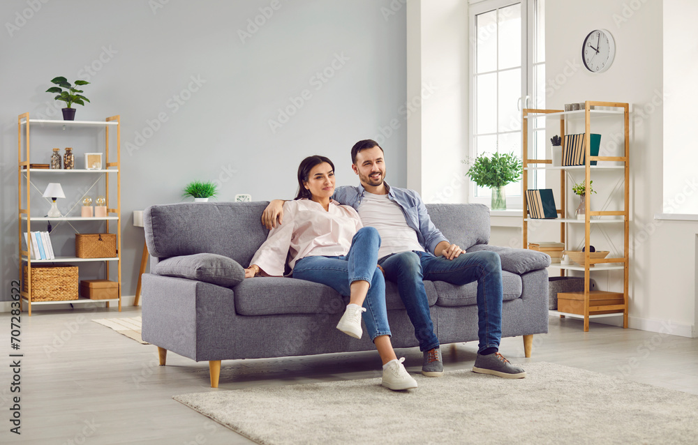 Couple watching TV together while sitting on couch in living room. Cheerful spouses sitting hugging on comfy sofa watching television. Happy family spending time together at home Couple watching TV together while sitting on couch in living room. Cheerful spouses sitting hugging on comfy sofa watching television. Happy family spending time together at home