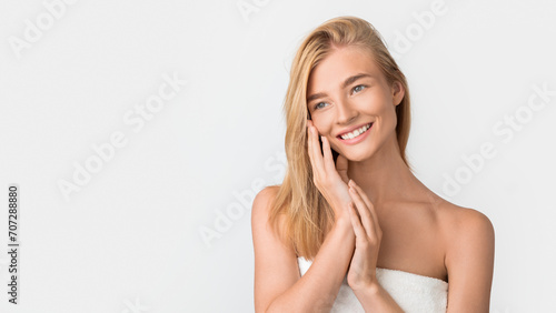 Beautiful blonde woman wrapped in towel touches face, white studio