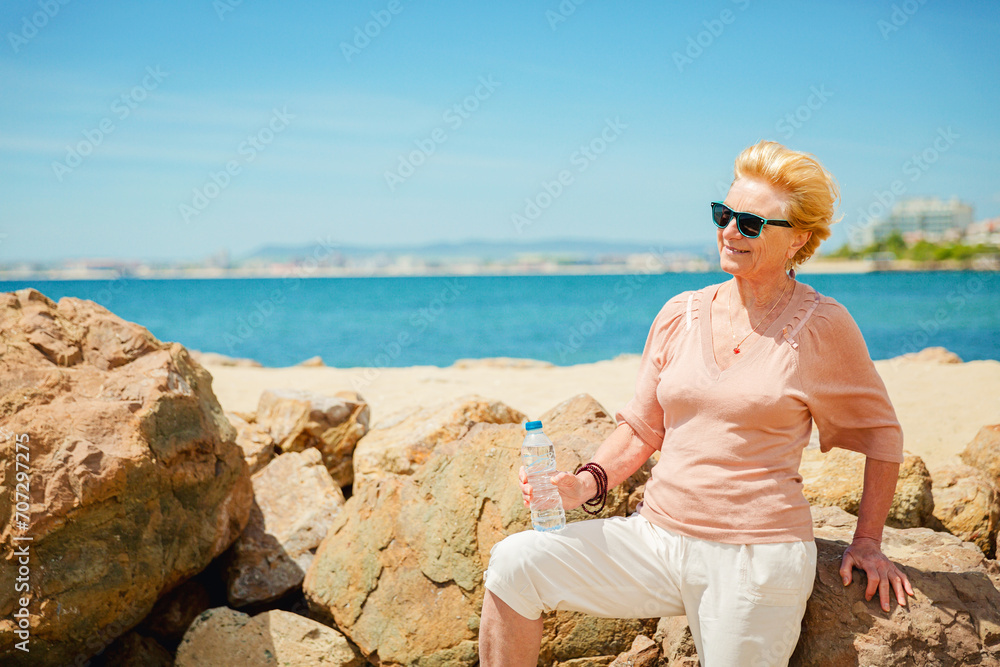 Retro style portrait of attractive senior woman sitting on the beach with a bottle of water wearing dark sunglasses. Old woman enjoying life