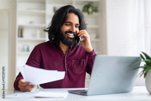 Indian man discussing work on phone while reviewing documents and using laptop photo