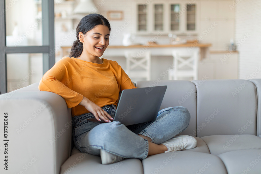 Smiling young indian woman using her laptop while sitting cross-legged on couch Smiling young indian woman using her laptop while sitting cross-legged on couch