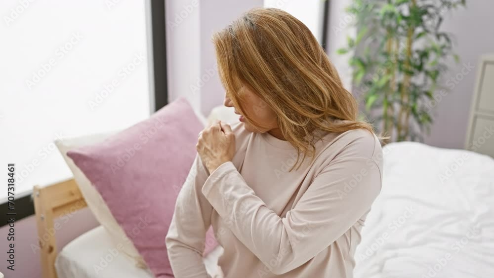 A woman expressing pain while clutching her shoulder in a bedroom setting. A woman expressing pain while clutching her shoulder in a bedroom setting.