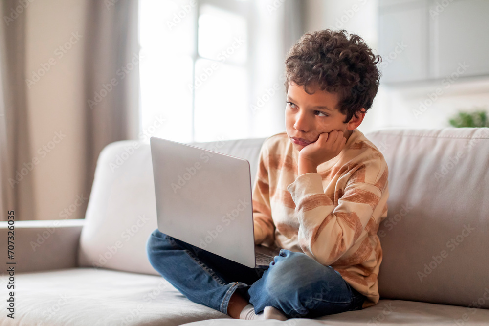 Pensive little black boy sitting with laptop on couch and looking away Pensive little black boy sitting with laptop on couch and looking away