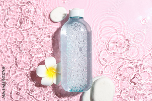 Bottle of micellar water with spa stones and plumeria flower in water on pink background