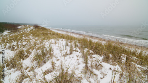 Morze Bałtyckie zimą. Śnieg pokrywa wydmy i rudawozłotą trawę. Mglisty horyzont. Długi czas naświetlania. Rozmyte fale, wyraziste kamyki.