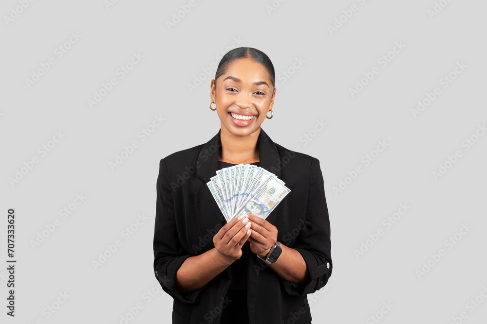 Smiling African American businesswoman in a stylish black blazer joyfully holding a fan of cash Smiling African American businesswoman in a stylish black blazer joyfully holding a fan of cash