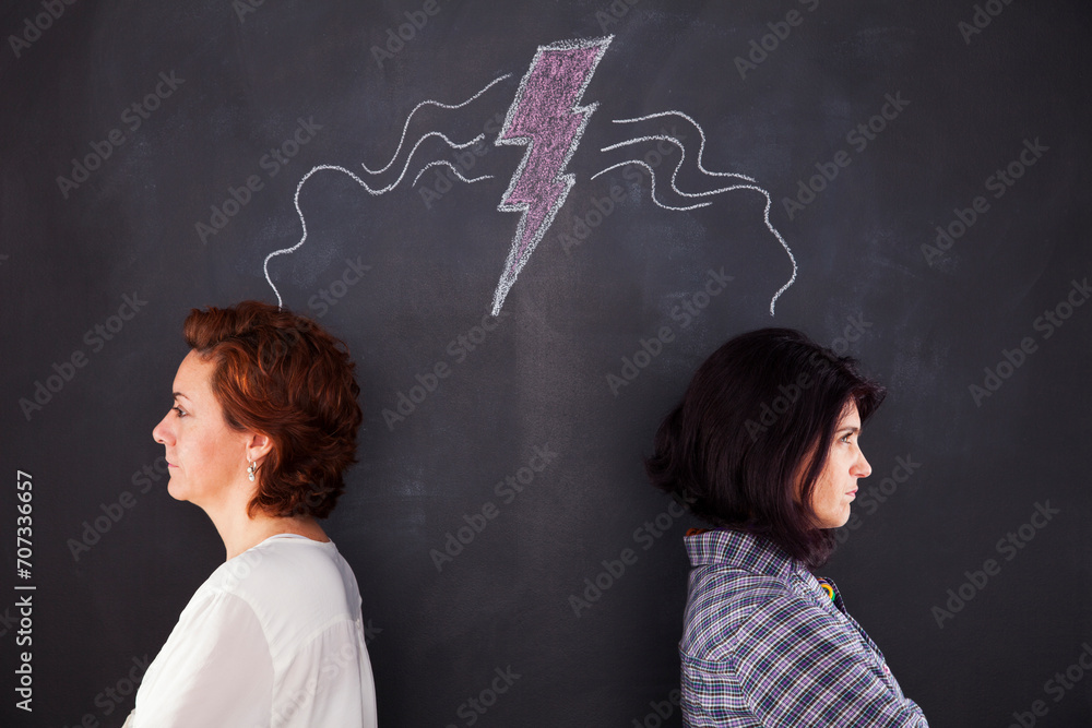Two women stand by a chalkboard, their backs turned to each other, exuding an air of discontent. In this tense scene, the chalkboard becomes a silent witness to a moment of discord