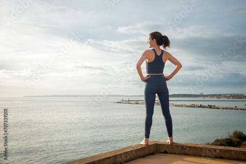 Rear view of a sporty woman facing ocean with hands on hips, relaxing after exercise.