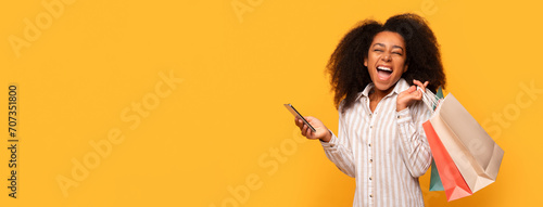 Happy black shopper with bags using phone on yellow background, panorama with free space photo