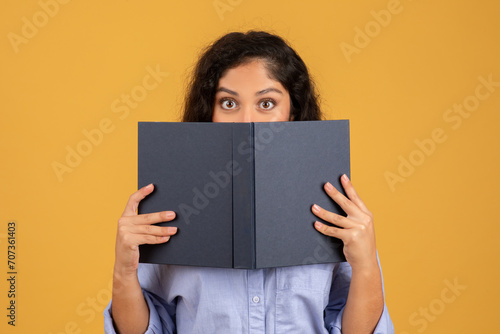 Surprised young woman with curly hair peeking over an open dark book photo