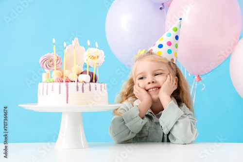 Cute little girl in party hat with balloons and Birthday cake on table against blue background